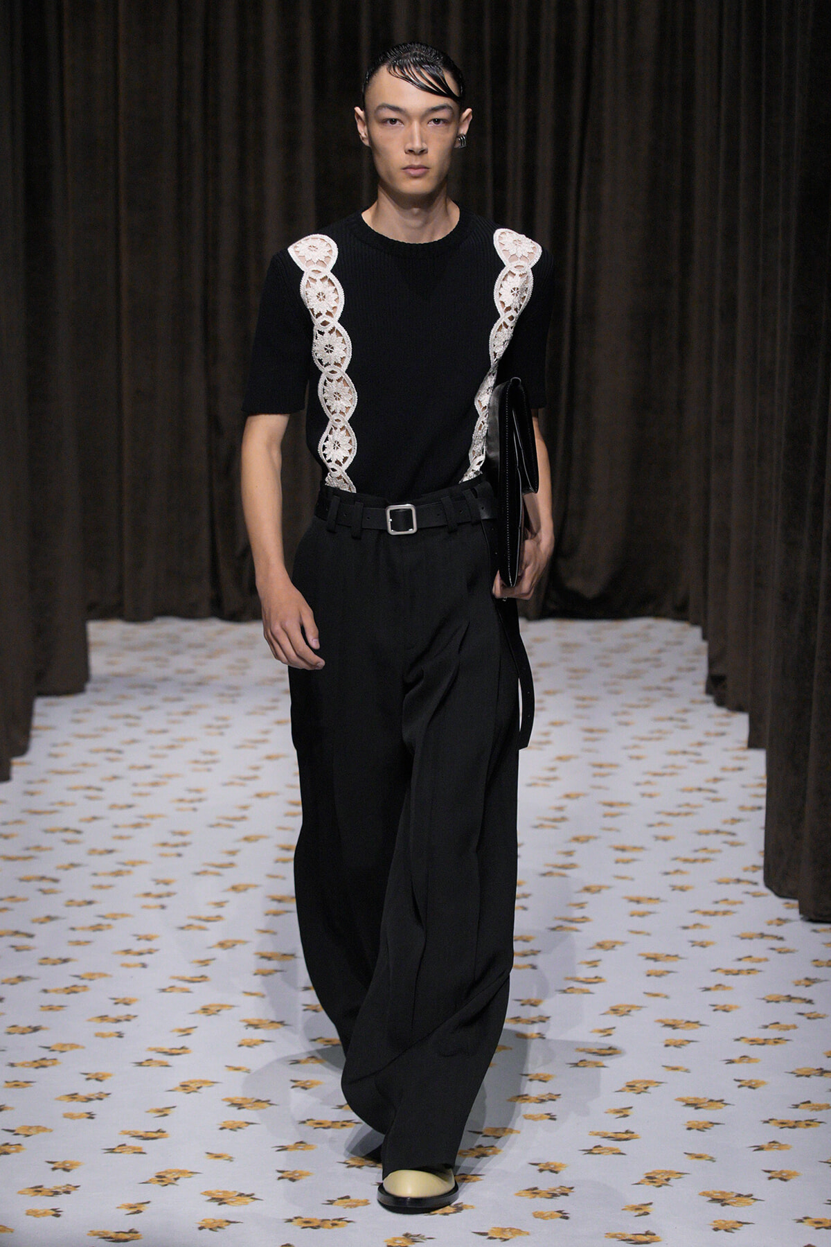 Male model on a fashion-show runway wearing a black top with white lace panels down the sides, black wide-leg trousers, and a black clutch/binder tucked under the arm.