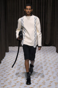 Male model walks a runway wearing a cream long-sleeve tunic with white scalloped lace trim on the sides, black shorts, and black boots, carrying a black bag