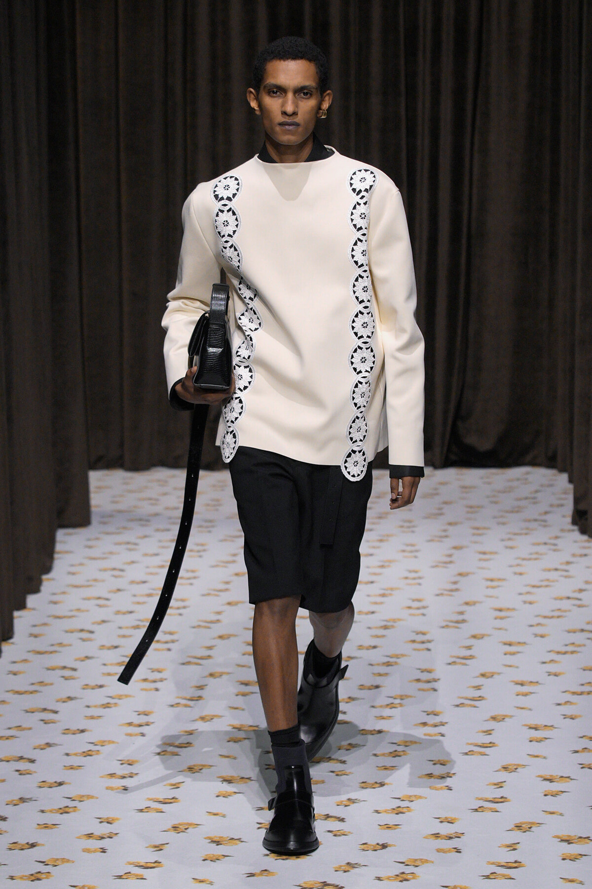 Male model walks a runway wearing a cream long-sleeve tunic with white scalloped lace trim on the sides, black shorts, and black boots, carrying a black bag