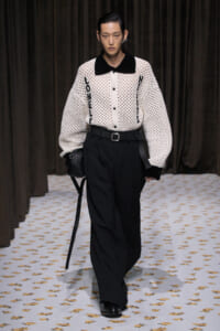 Runway model wearing a white open-knit cardigan with a black collar, black wide-leg pants, a belt, and a small black shoulder bag.