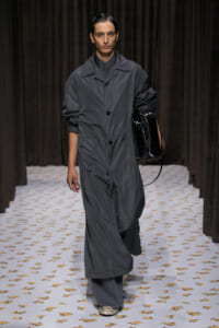 Model walks a runway in a loose gray coat with black buttons, carrying a glossy black handbag against a dark backdrop.