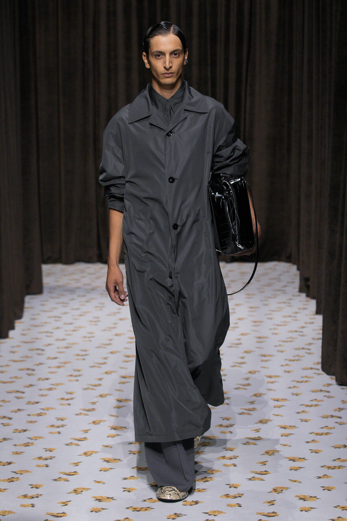 Model walks a runway in a loose gray coat with black buttons, carrying a glossy black handbag against a dark backdrop.