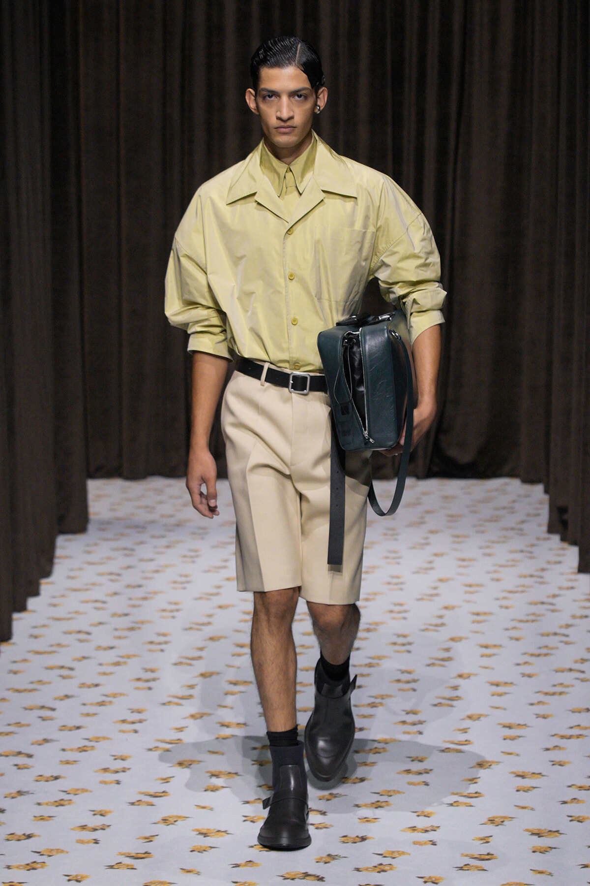 Male model on a runway wearing an oversized pale yellow shirt, beige shorts, black belt, and carrying a dark green bag under his arm.