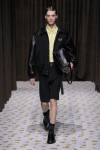 Male model walks a runway in a black jacket with leather sleeves, pale yellow shirt, black shorts, and black boots, carrying a black bag.