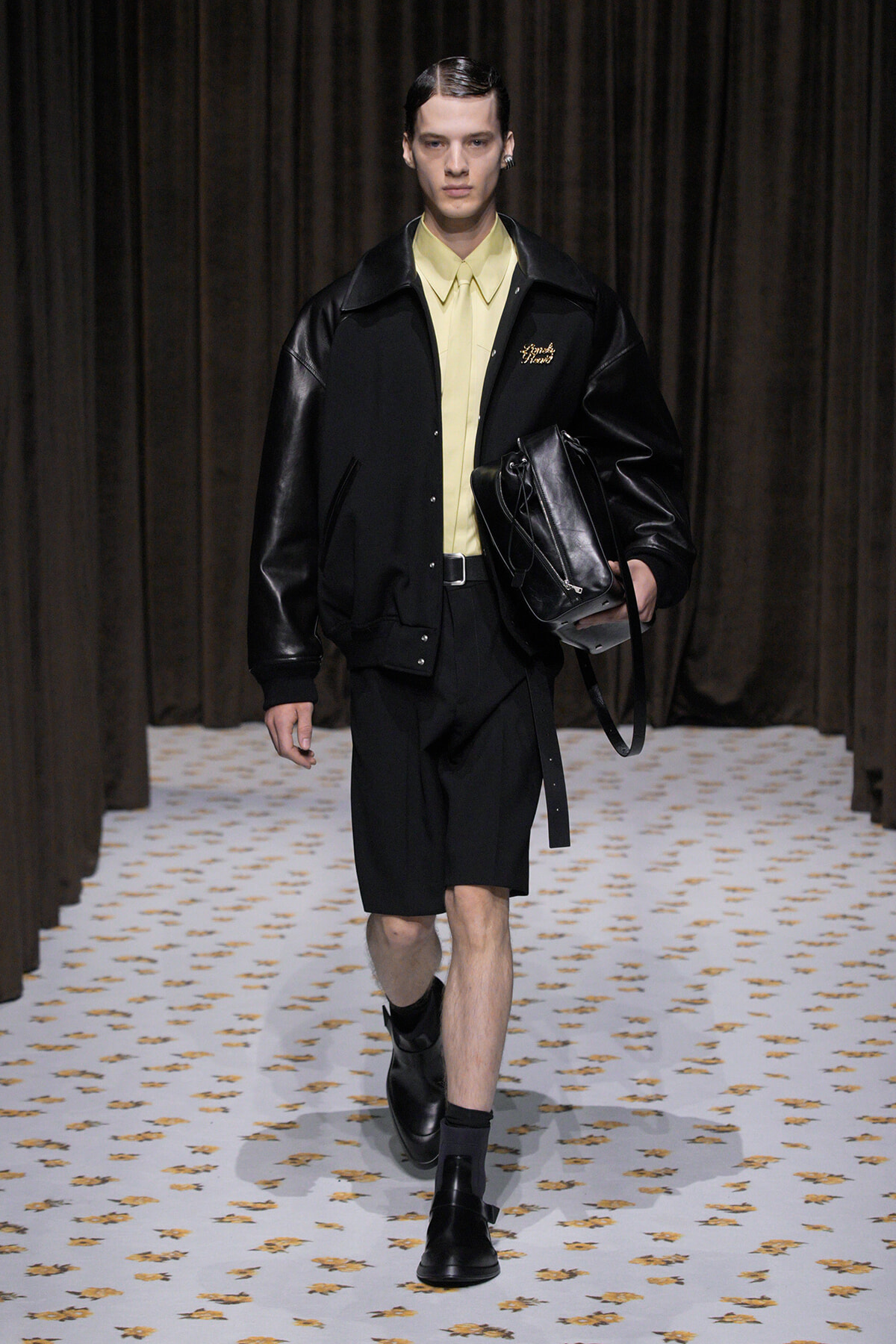 Male model walks a runway in a black jacket with leather sleeves, pale yellow shirt, black shorts, and black boots, carrying a black bag.