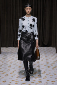 Runway model wearing a light blue cardigan with black floral embroidery, a black leather midi skirt, belt, and tall black boots, carrying a brown handbag.