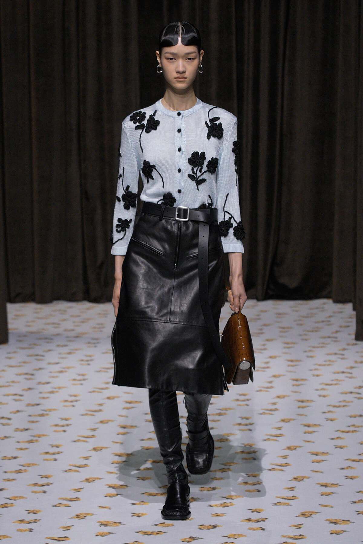 Runway model wearing a light blue cardigan with black floral embroidery, a black leather midi skirt, belt, and tall black boots, carrying a brown handbag.