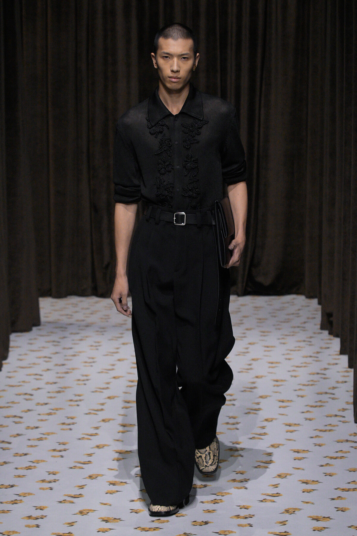 Male model walking a fashion runway in all-black ensemble with embroidered top, wide trousers, belt, and snakeskin shoes, carrying a black clutch