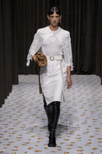 Runway model in an all-white look: white sweater and skirt with a floral-embellished cape, paired with a white belt and black over-the-knee boots, holding a tan clutch.