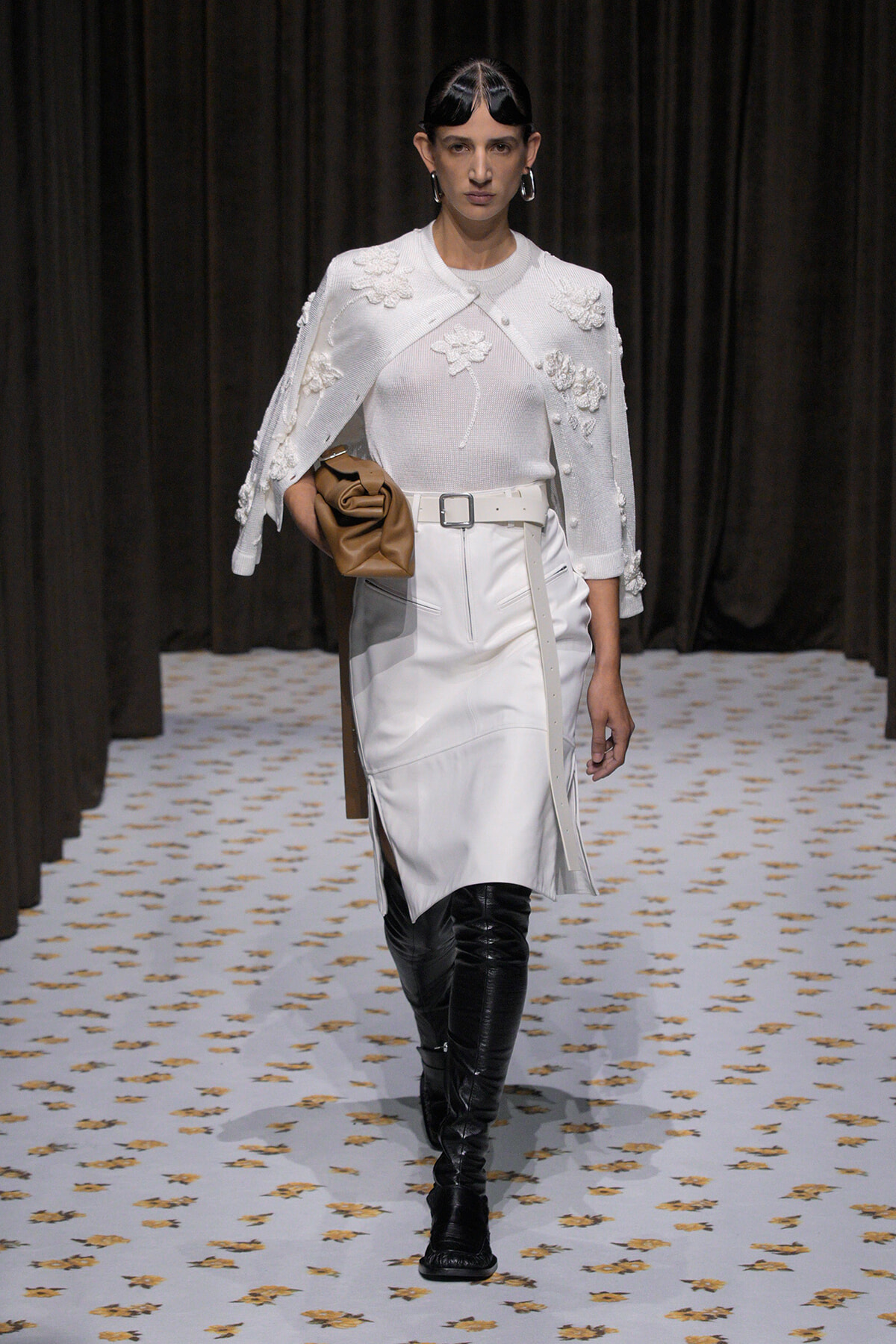 Runway model in an all-white look: white sweater and skirt with a floral-embellished cape, paired with a white belt and black over-the-knee boots, holding a tan clutch.