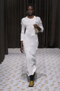Model walks a white, floral-embellished knit dress down a runway, carrying a small patterned handbag.