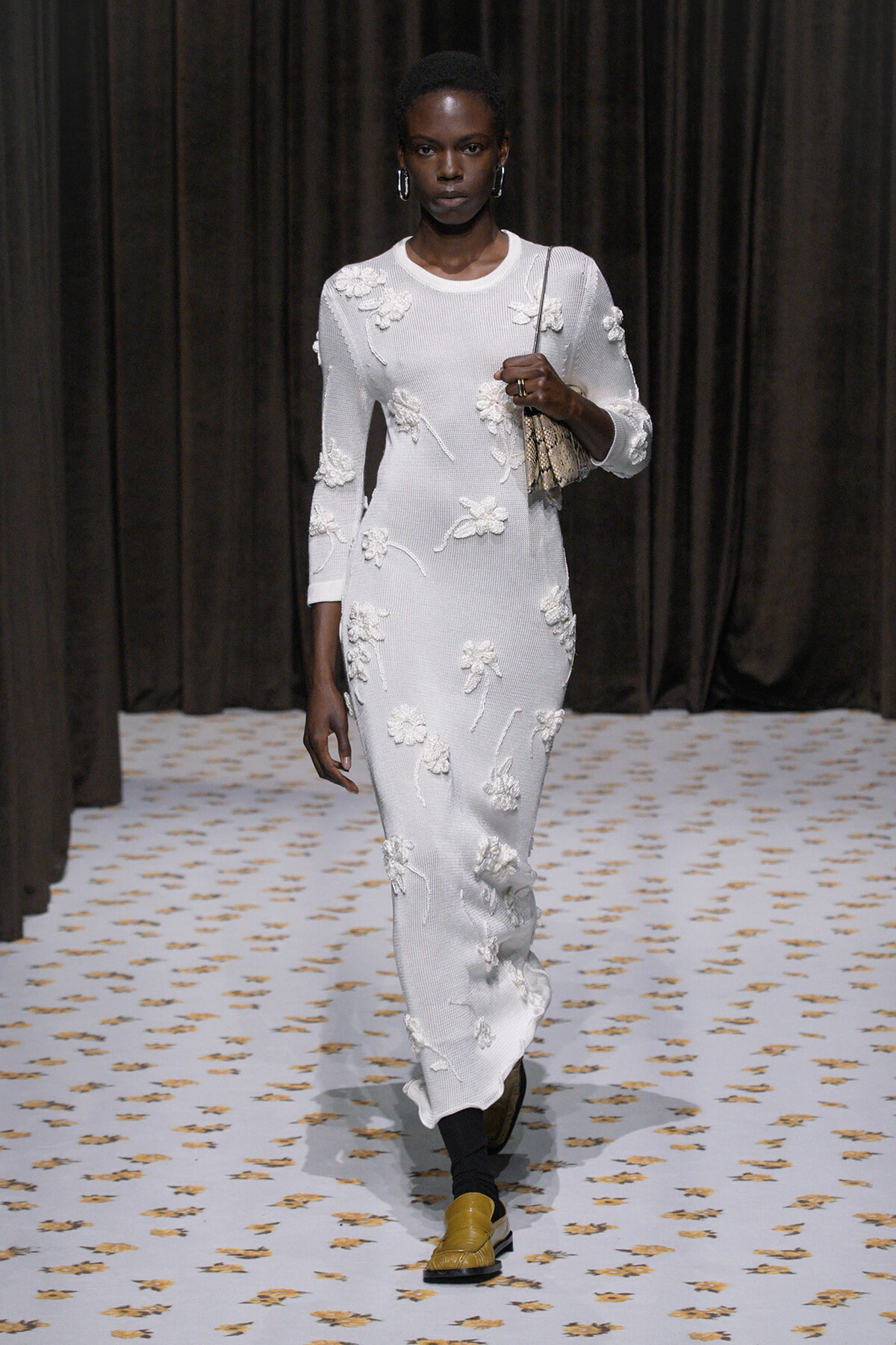 Model walks a white, floral-embellished knit dress down a runway, carrying a small patterned handbag.