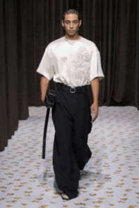 Male model in a white oversized top with raised embroidery and black wide-leg trousers, belt, and a black clutch on a runway.