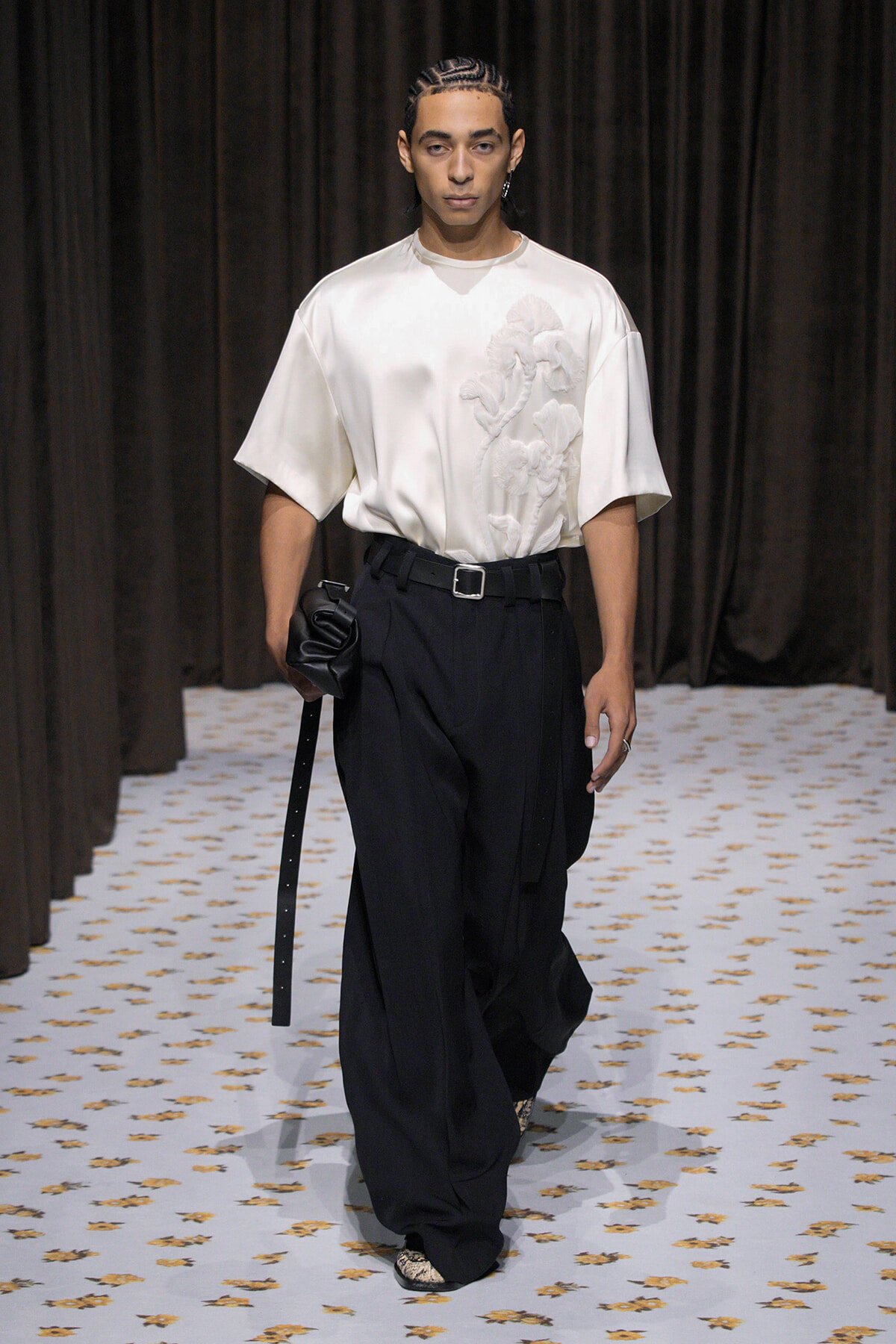 Male model in a white oversized top with raised embroidery and black wide-leg trousers, belt, and a black clutch on a runway.