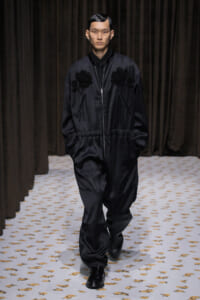 Male model walks a runway in a black satin bomber jacket with floral appliqués, matching pants, and black shoes; hands in pockets, neutral expression.