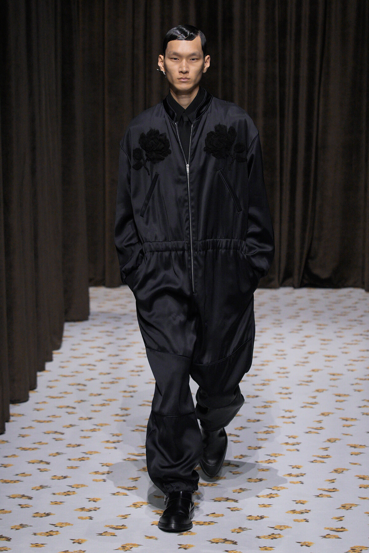 Male model walks a runway in a black satin bomber jacket with floral appliqués, matching pants, and black shoes; hands in pockets, neutral expression.