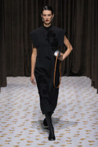 Female model walks a fashion runway in a black dress with floral embroidery, carrying a small round silver bag with a tan strap.