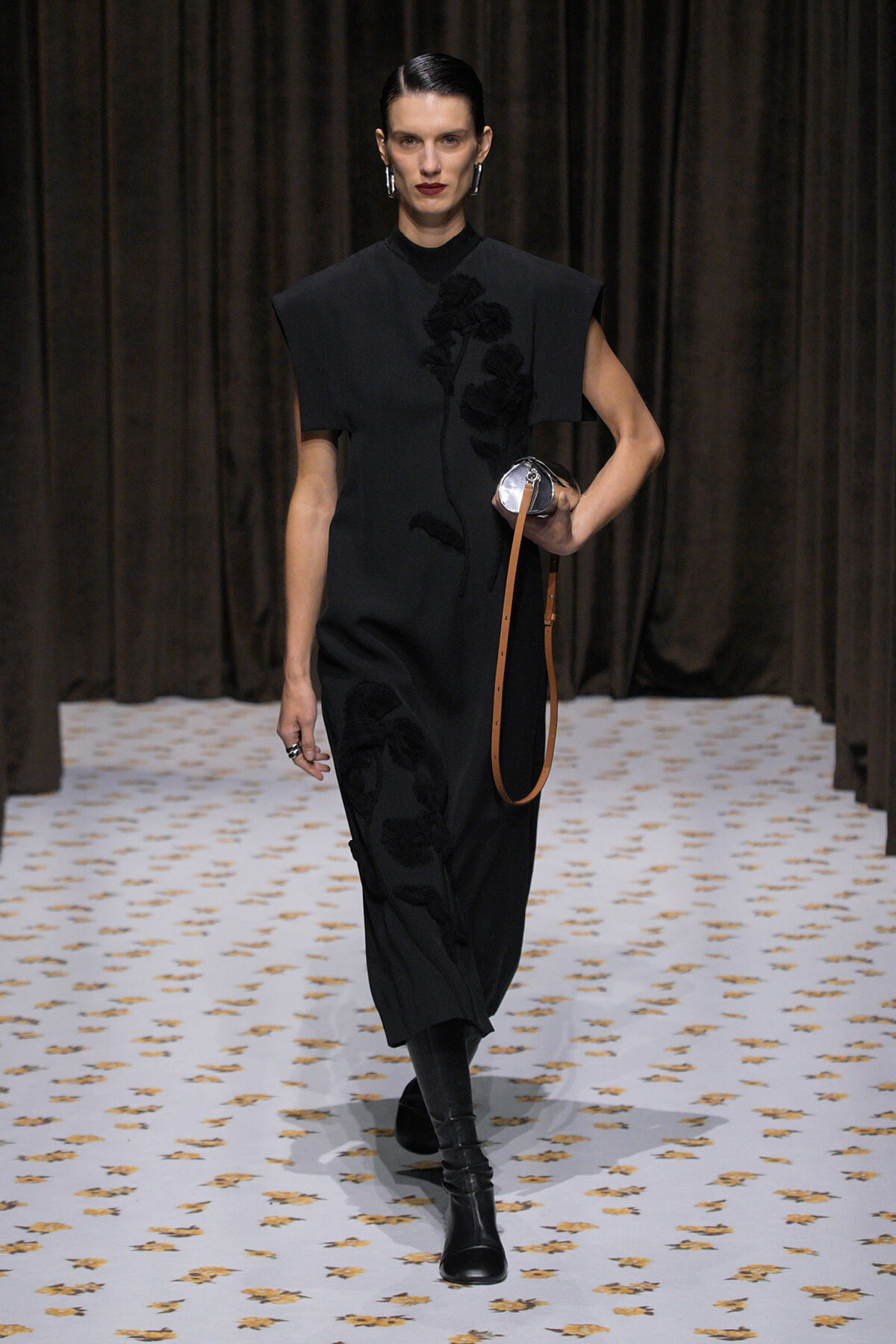 Female model walks a fashion runway in a black dress with floral embroidery, carrying a small round silver bag with a tan strap.