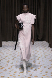 Model walks a runway in a pale pink dress with white floral appliqué, white knee-high boots, and a round black handbag.