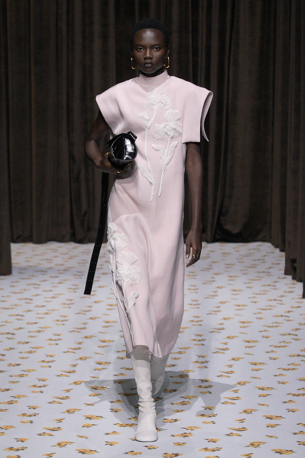Model walks a runway in a pale pink dress with white floral appliqué, white knee-high boots, and a round black handbag.
