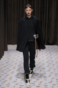 Model walks a fashion runway in an all-black ensemble: oversized blazer over a ribbed black top, matching ribbed pants with silver trim, and white heels with black socks; holds a tan strap accessory.