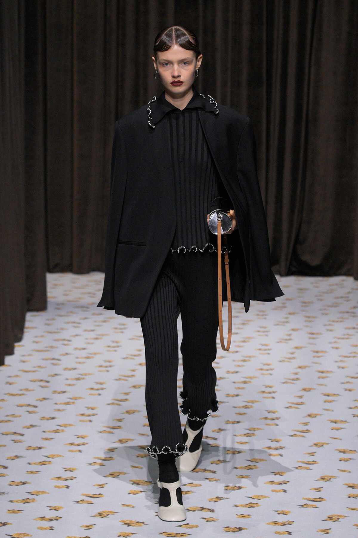 Model walks a fashion runway in an all-black ensemble: oversized blazer over a ribbed black top, matching ribbed pants with silver trim, and white heels with black socks; holds a tan strap accessory.