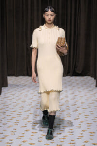 Runway model in a cream ribbed dress with pearl trims, carrying a brown studded clutch.
