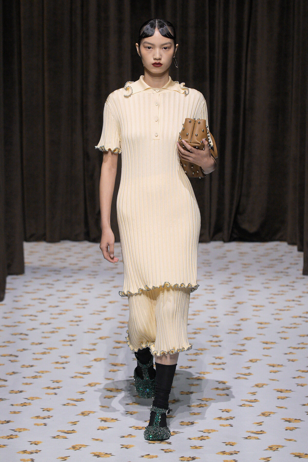 Runway model in a cream ribbed dress with pearl trims, carrying a brown studded clutch.