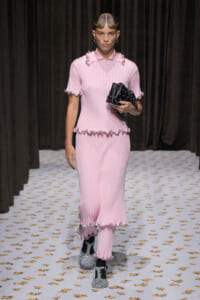 Model walks the runway in a pink ribbed knit dress with scalloped trim, holding a black studded handbag.