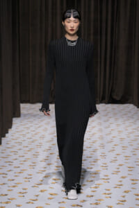 Model walking a runway in a black vertically ribbed full-length dress with scalloped cuffs and white shoes.