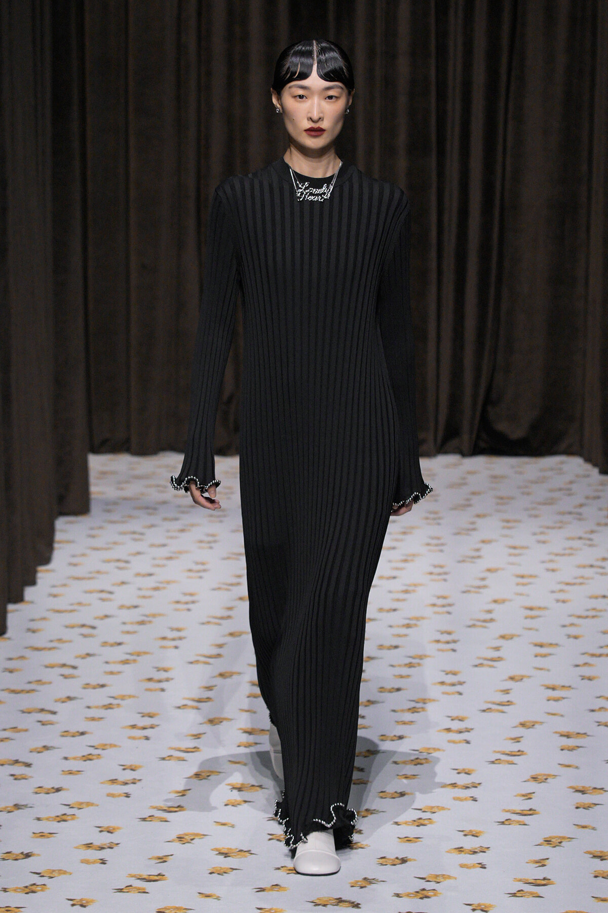 Model walking a runway in a black vertically ribbed full-length dress with scalloped cuffs and white shoes.