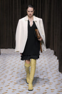 Model walking the runway in a white oversized blazer over a black mini dress with ruffle hem, paired with tall yellow knee-high boots and a brown clutch tucked under her arm.