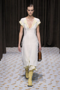 Model walks a runway in a white, sheer dress with pale yellow ruffle accents and matching yellow knee-high boots, carrying a brown handbag.