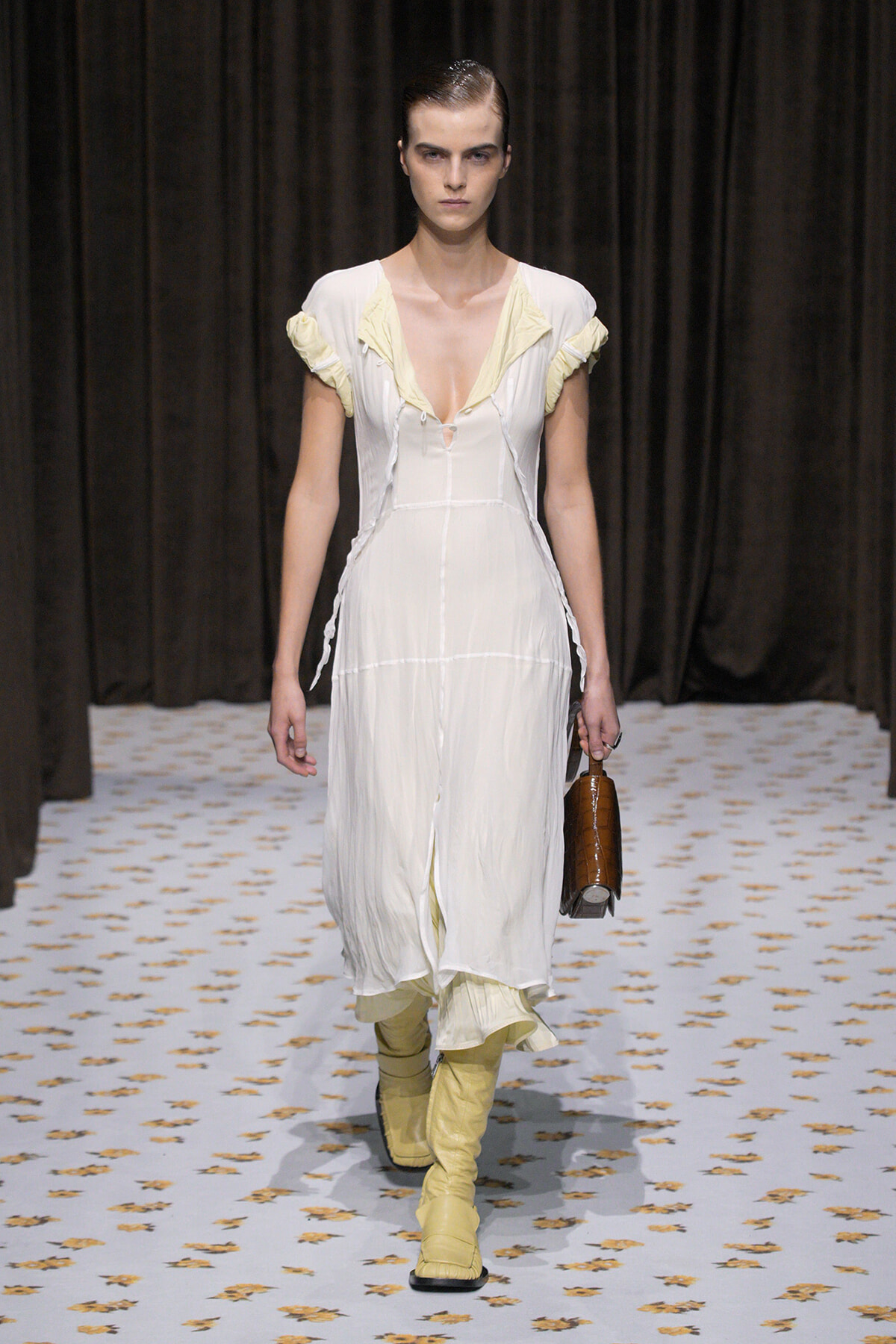 Model walks a runway in a white, sheer dress with pale yellow ruffle accents and matching yellow knee-high boots, carrying a brown handbag.