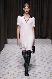 Model walking a fashion runway in a white knee-length dress with black collar, ruffled sleeves, and black thigh-high boots, carrying a small black handbag.
