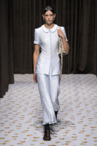 Model walks runway in pale blue floral suit with a Peter Pan collar and matching wide-leg pants, carrying a snakeskin bag over the shoulder.