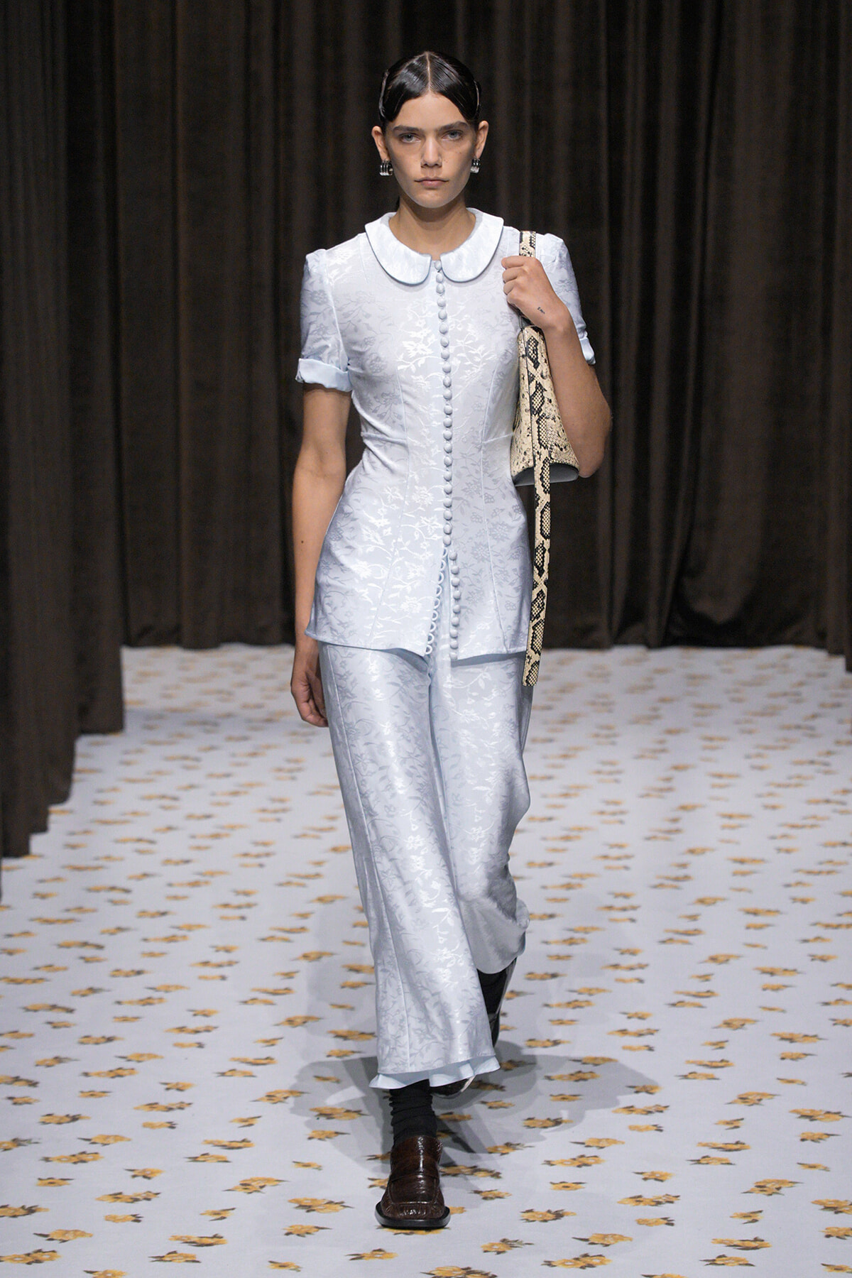 Model walks runway in pale blue floral suit with a Peter Pan collar and matching wide-leg pants, carrying a snakeskin bag over the shoulder.