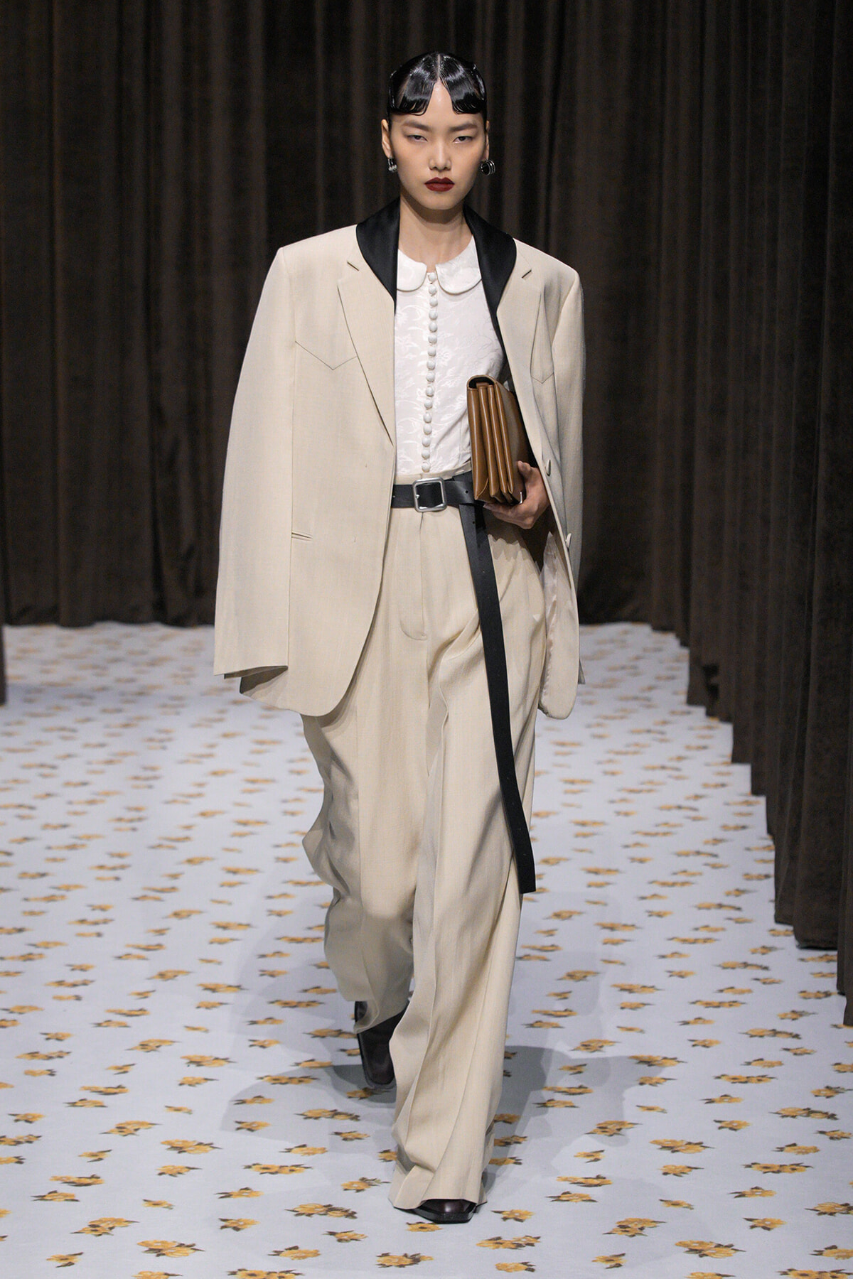 Model walks a runway in a beige oversized suit with black satin lapels, white blouse, and matching trousers, holding a brown clutch.