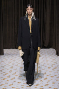 Model walking runway in oversized black suit over tan top, white fringe earrings, carrying tan handbag against a brown curtain backdrop.