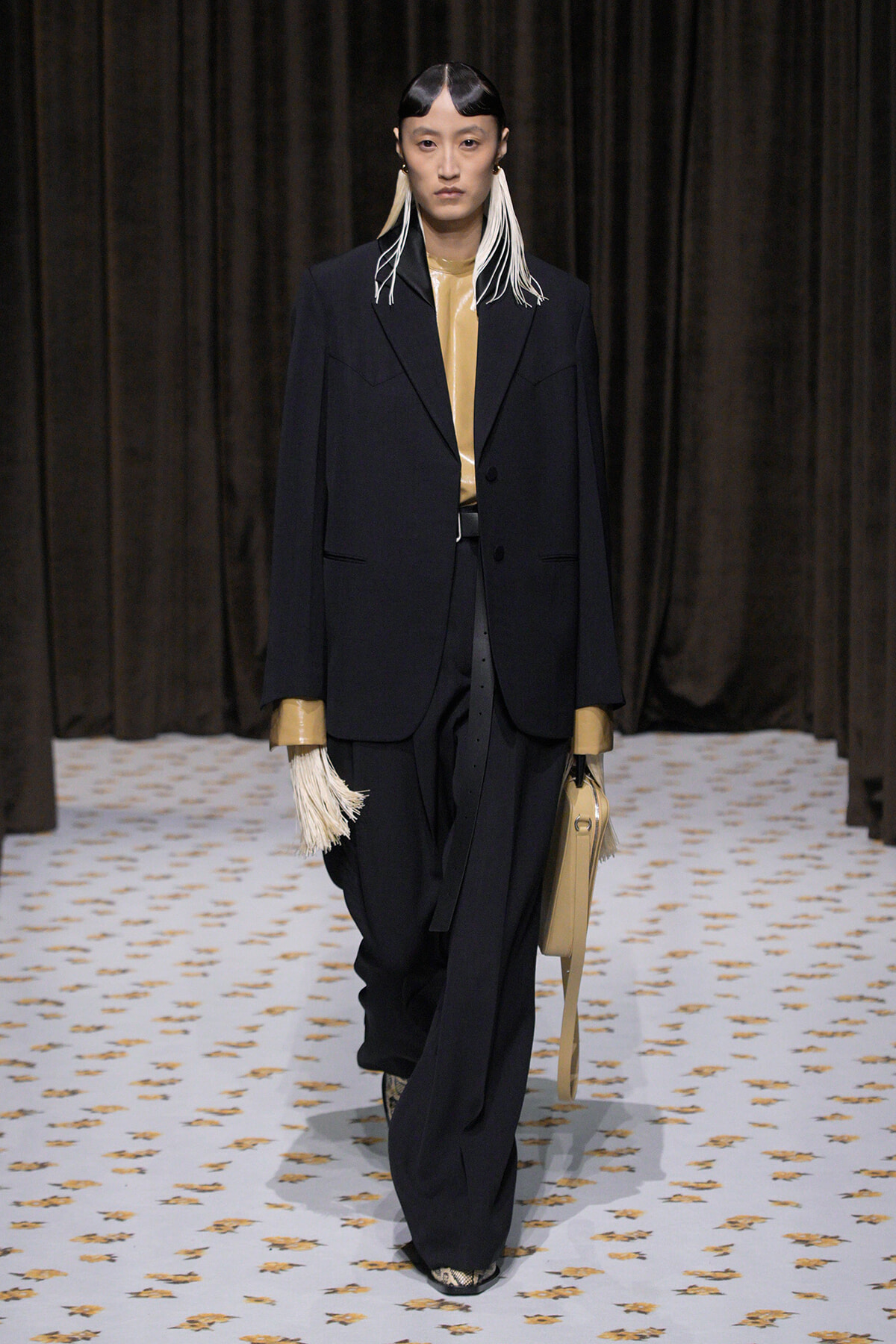 Model walking runway in oversized black suit over tan top, white fringe earrings, carrying tan handbag against a brown curtain backdrop.