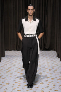 Male model walking a runway in a black-and-white outfit: white center shirt panels with black sleeves, black high-waisted trousers, and a white belt.