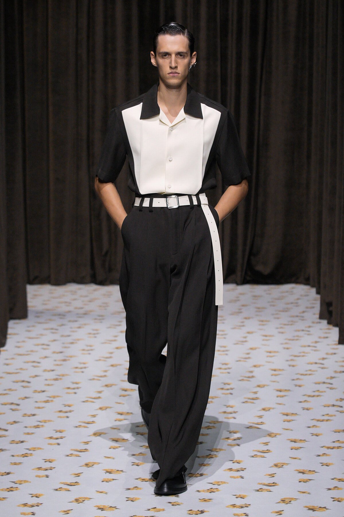 Male model walking a runway in a black-and-white outfit: white center shirt panels with black sleeves, black high-waisted trousers, and a white belt.
