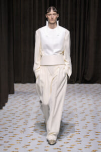 Model walking the runway in an ivory, structured top with a wide belt and matching wide-leg pants; snakeskin shoes.