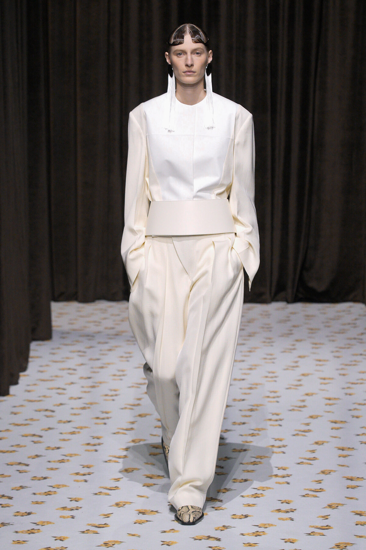 Model walking the runway in an ivory, structured top with a wide belt and matching wide-leg pants; snakeskin shoes.