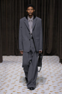 Model walks the runway in an oversized gray suit with a matching shirt, holding a black handbag against a dark curtain backdrop.