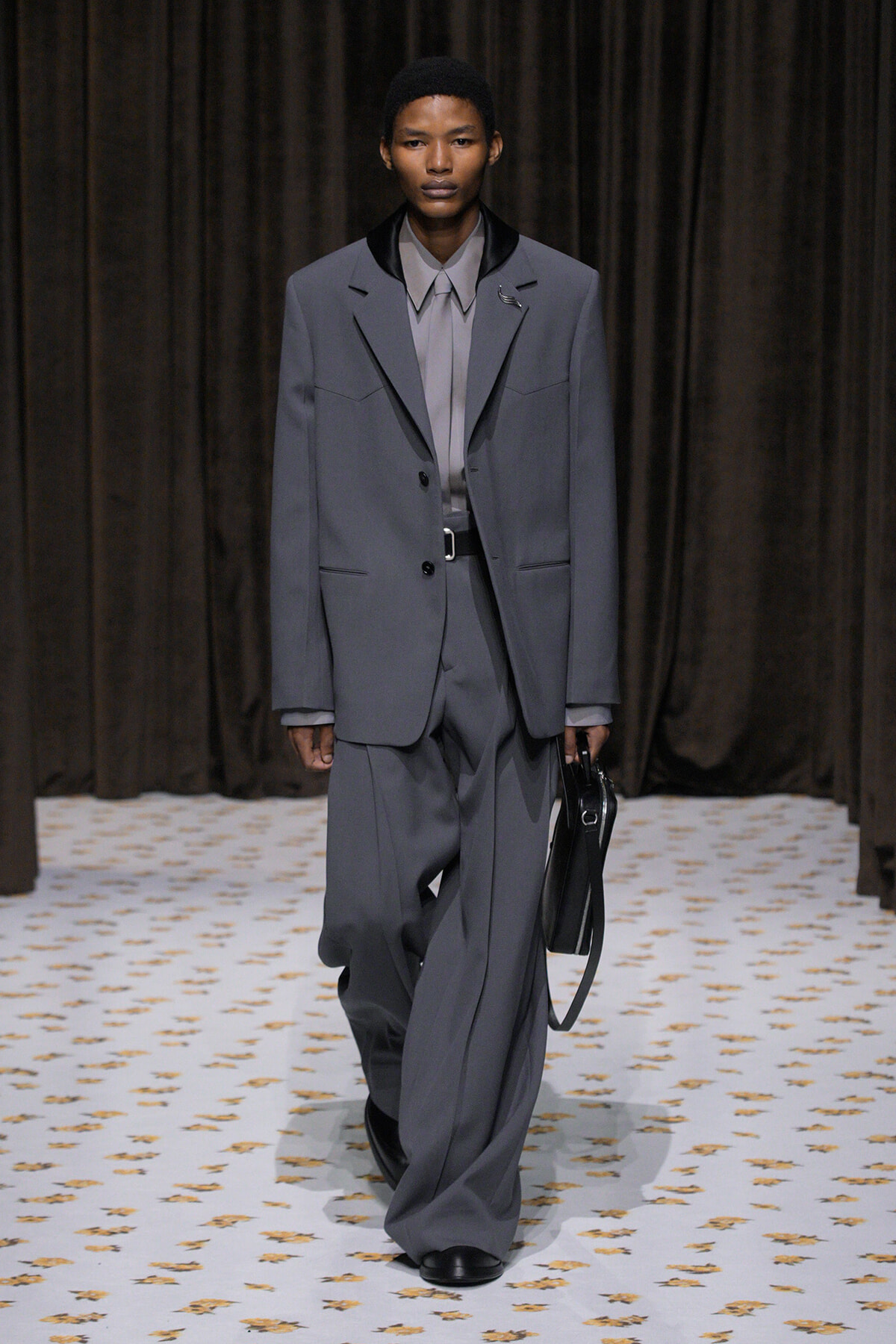 Model walks the runway in an oversized gray suit with a matching shirt, holding a black handbag against a dark curtain backdrop.