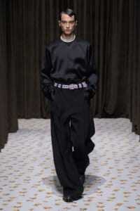 Male model walks a fashion show wearing a black satin top with high-contrast striped collar, wide-leg black trousers, and a lavender woven belt; hands in pockets.