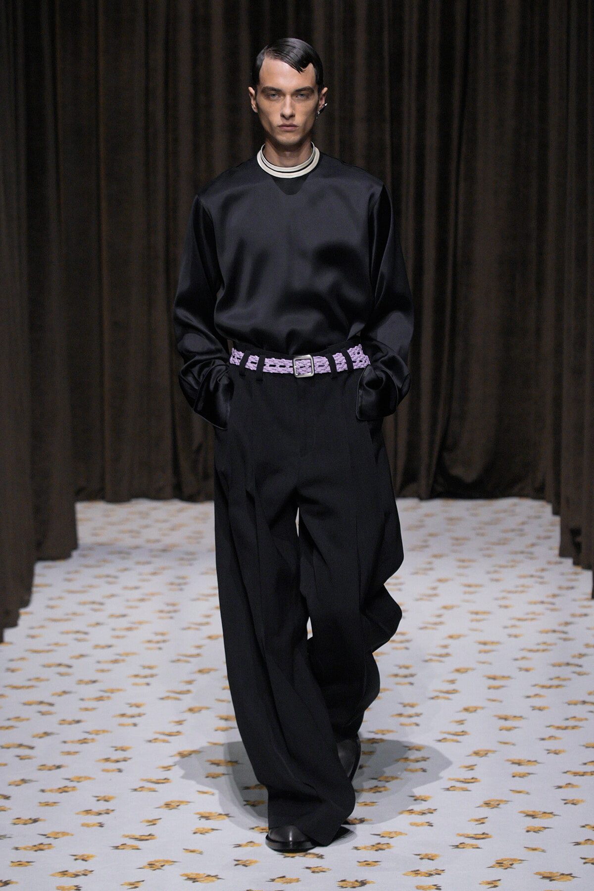 Male model walks a fashion show wearing a black satin top with high-contrast striped collar, wide-leg black trousers, and a lavender woven belt; hands in pockets.