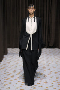 Model on a runway wearing a loose black suit with a white bib-style front and fringe details at the sleeves and gloves.