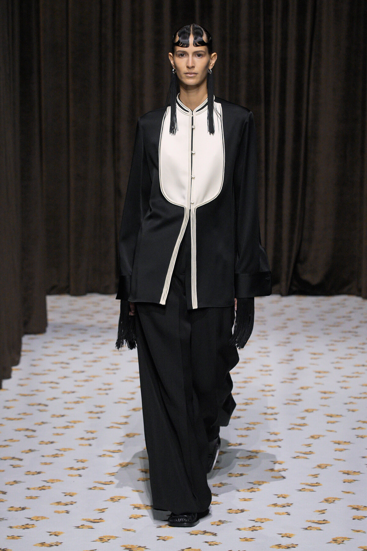 Model on a runway wearing a loose black suit with a white bib-style front and fringe details at the sleeves and gloves.